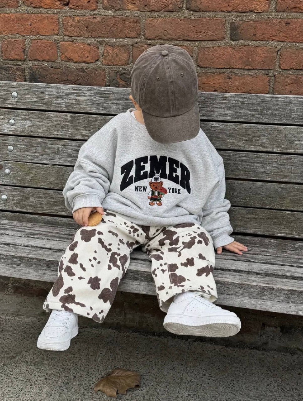 Young child seated on a bench in the heather grey ZEMER Bear Crewneck, three-quarter view of the varsity ZEMER arch and the bear holding red roses