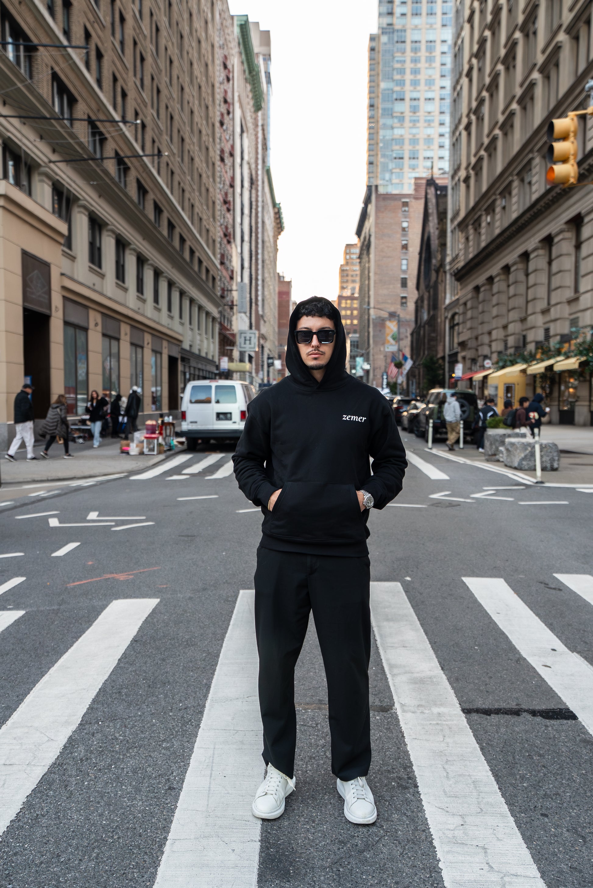 Person wearing a black hoodie and pants standing on a city street with tall buildings in the background.