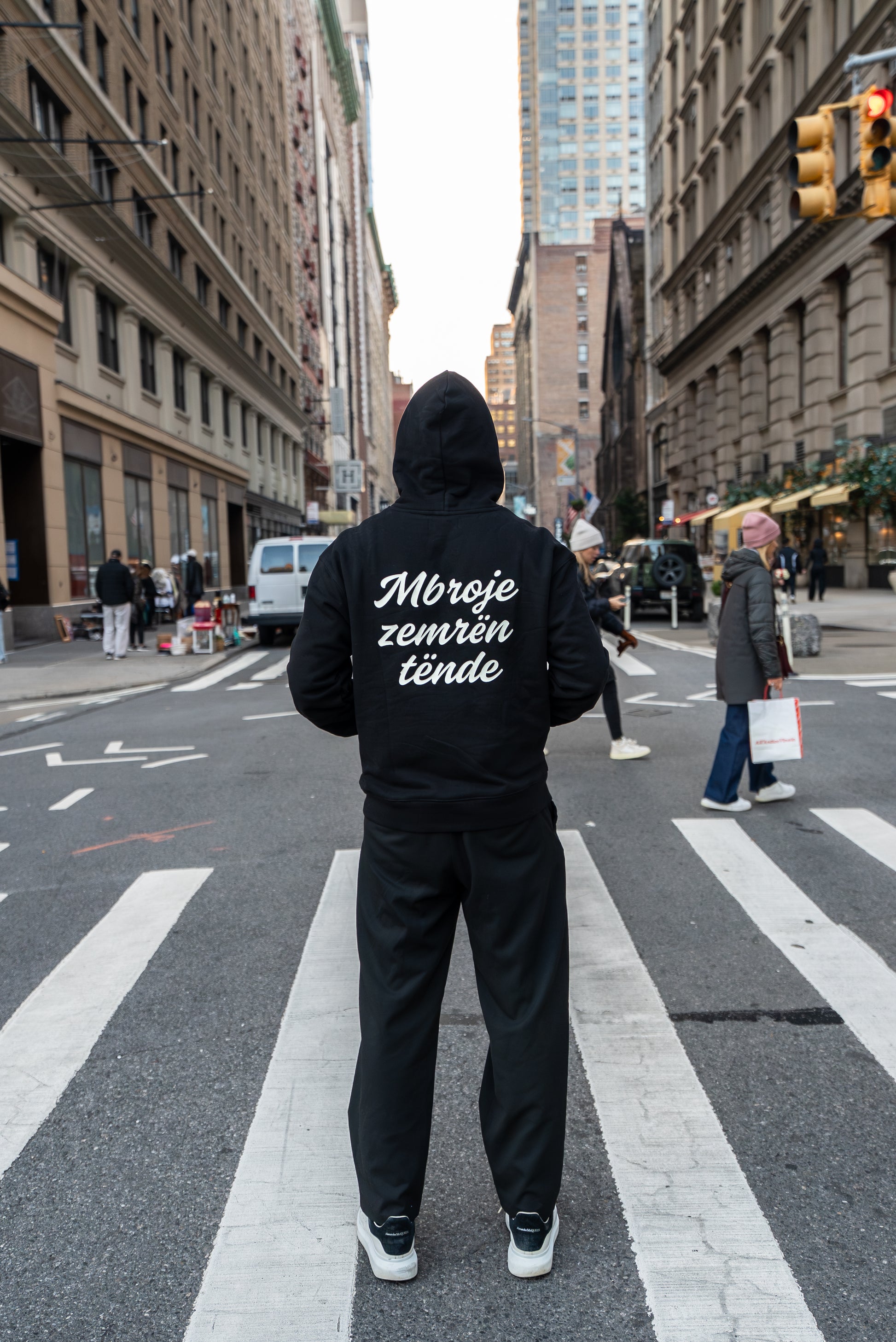Person wearing a black hoodie with text on the back, standing on a city street.