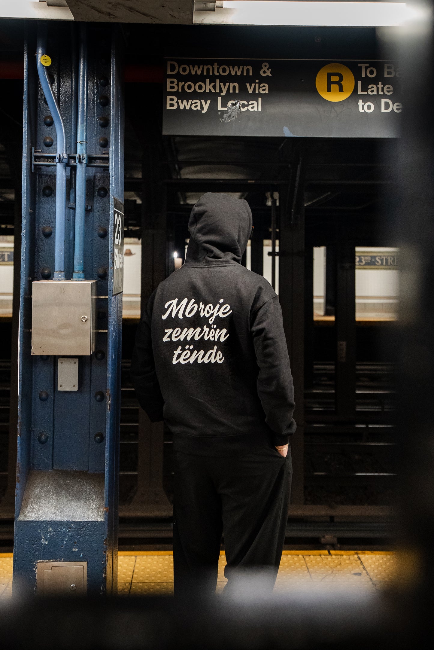 Person wearing a black hoodie with text on the back in a subway station.