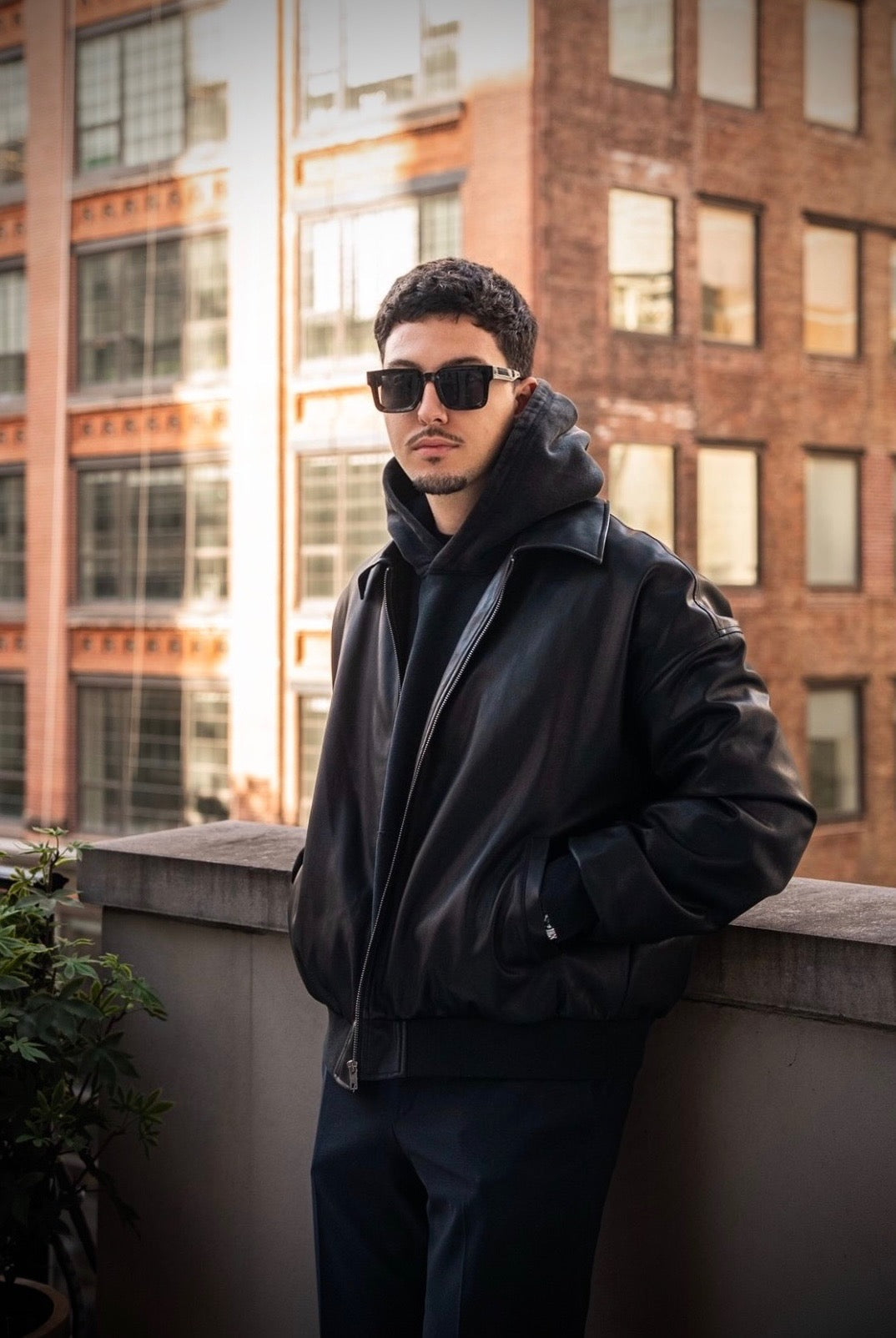 Man wearing sunglasses and a black jacket standing on a rooftop with city buildings in the background