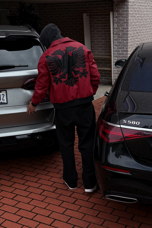 Person wearing a red jacket with black eagle design, standing between two cars on a brick driveway.