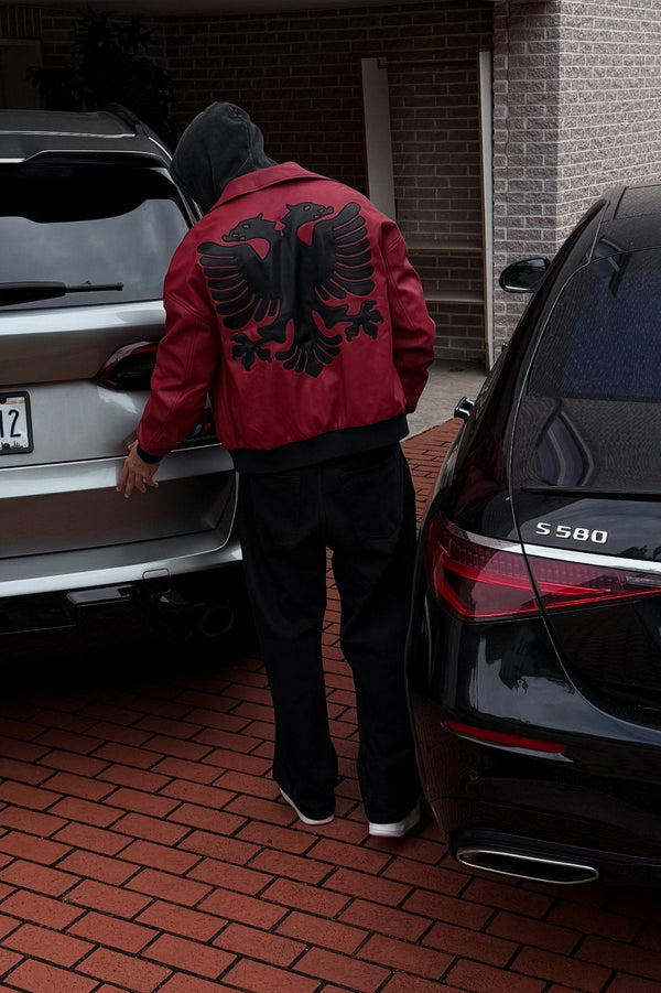 Person wearing a red jacket with black eagle design, standing between two cars on a brick driveway.