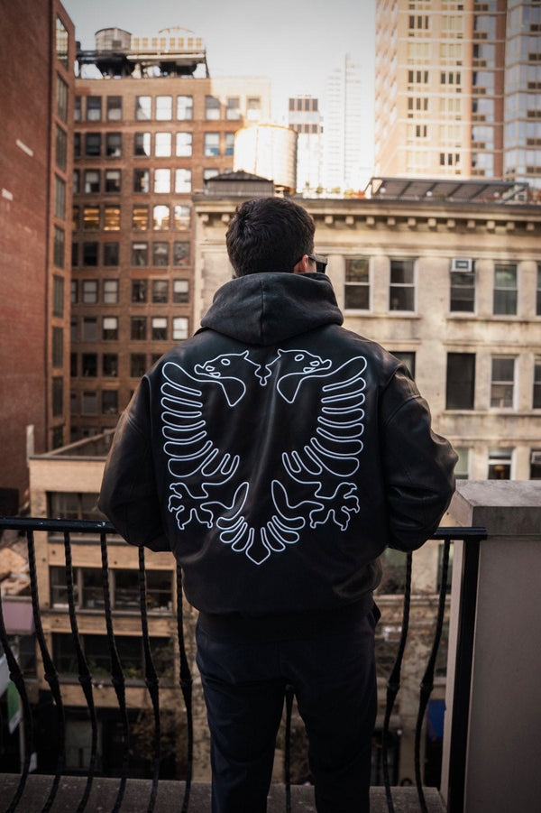 Person wearing a black hoodie with an eagle design on the back, standing on a rooftop with cityscape in the background.