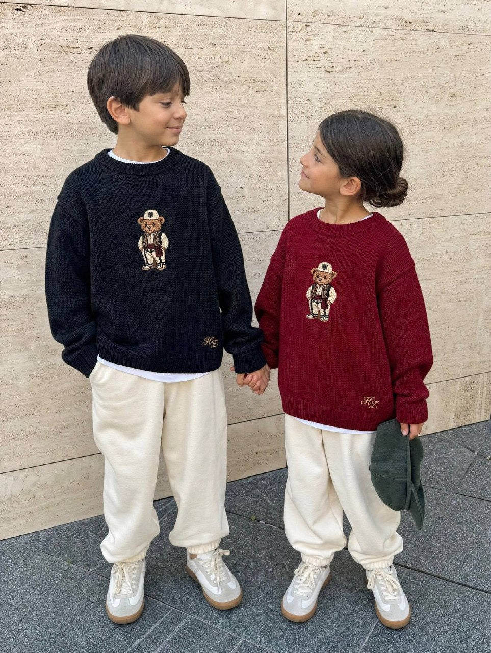 Albanian Ted Dua Bear Sweater for kids, a boy in black and a girl in burgundy holding hands against a travertine wall