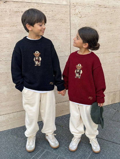 Albanian Ted Dua Bear Sweater for kids, a boy in black and a girl in burgundy holding hands against a travertine wall
