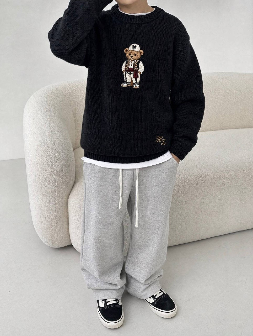 Albanian Ted Dua Bear Sweater for kids in black, a child beside a cream boucle sofa in grey sweatpants, hand raised to head