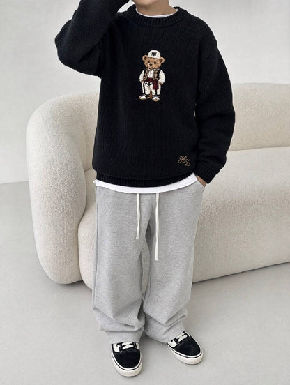 Albanian Ted Dua Bear Sweater for kids in black, a child beside a cream boucle sofa in grey sweatpants, hand raised to head