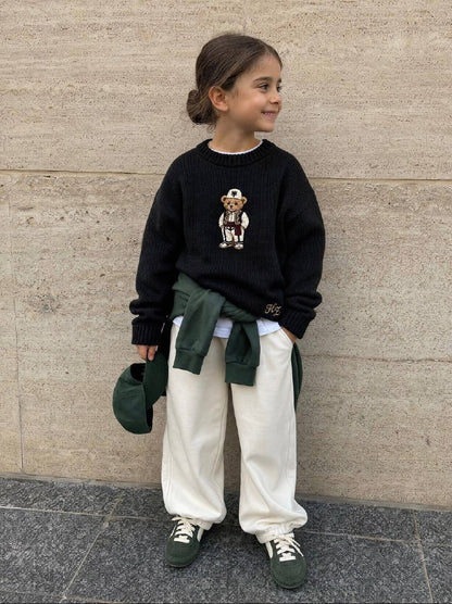 Albanian Ted Dua Bear Sweater for kids in black, a girl standing against a travertine wall with a green jacket at her waist