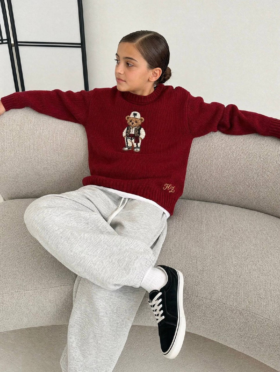 Albanian Ted Dua Bear Sweater for kids in burgundy, a girl seated on a cream boucle sofa with grey sweatpants