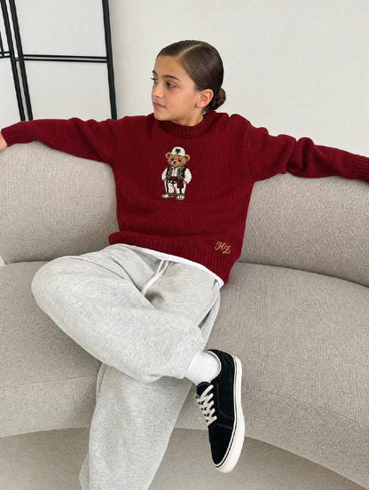 Albanian Ted Dua Bear Sweater for kids in burgundy, a girl seated on a cream boucle sofa with grey sweatpants