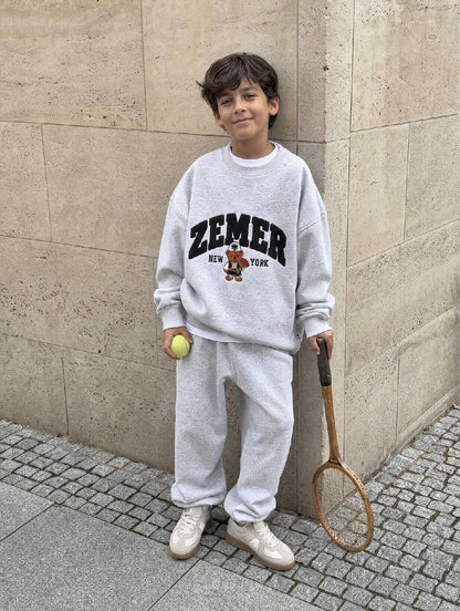 Boy in the heather grey ZEMER Bear Crewneck against a stone wall, full-length front view of the black varsity ZEMER arch and the bear in a qeleshe