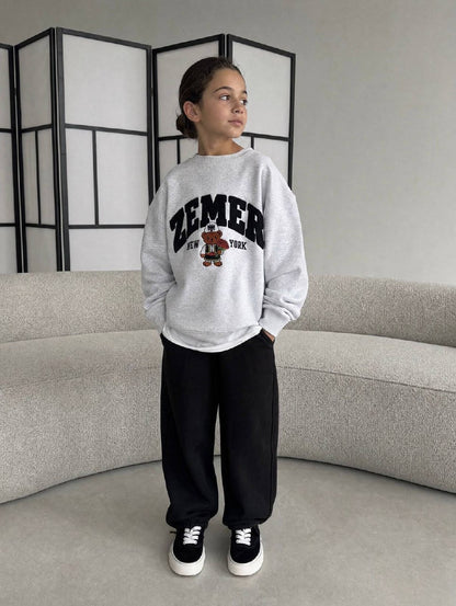 Girl in the heather grey ZEMER Bear Crewneck indoors, full-length front view of the oversized fit with the black varsity ZEMER arch over NEW YORK