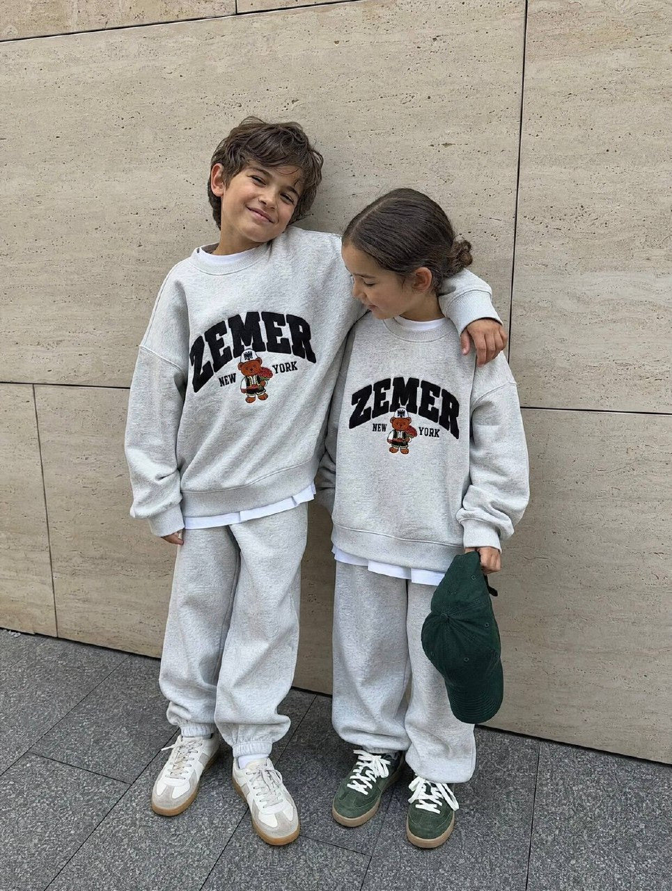 Two children in the heather grey ZEMER Bear Crewneck by a travertine wall, full-length front view of the black varsity ZEMER arch and embroidered bear